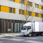 White box truck parked on the street in front of a modern building with yellow window panels.